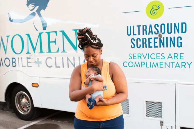 mom holding a newborn, standing in front of a mobile clinic bus